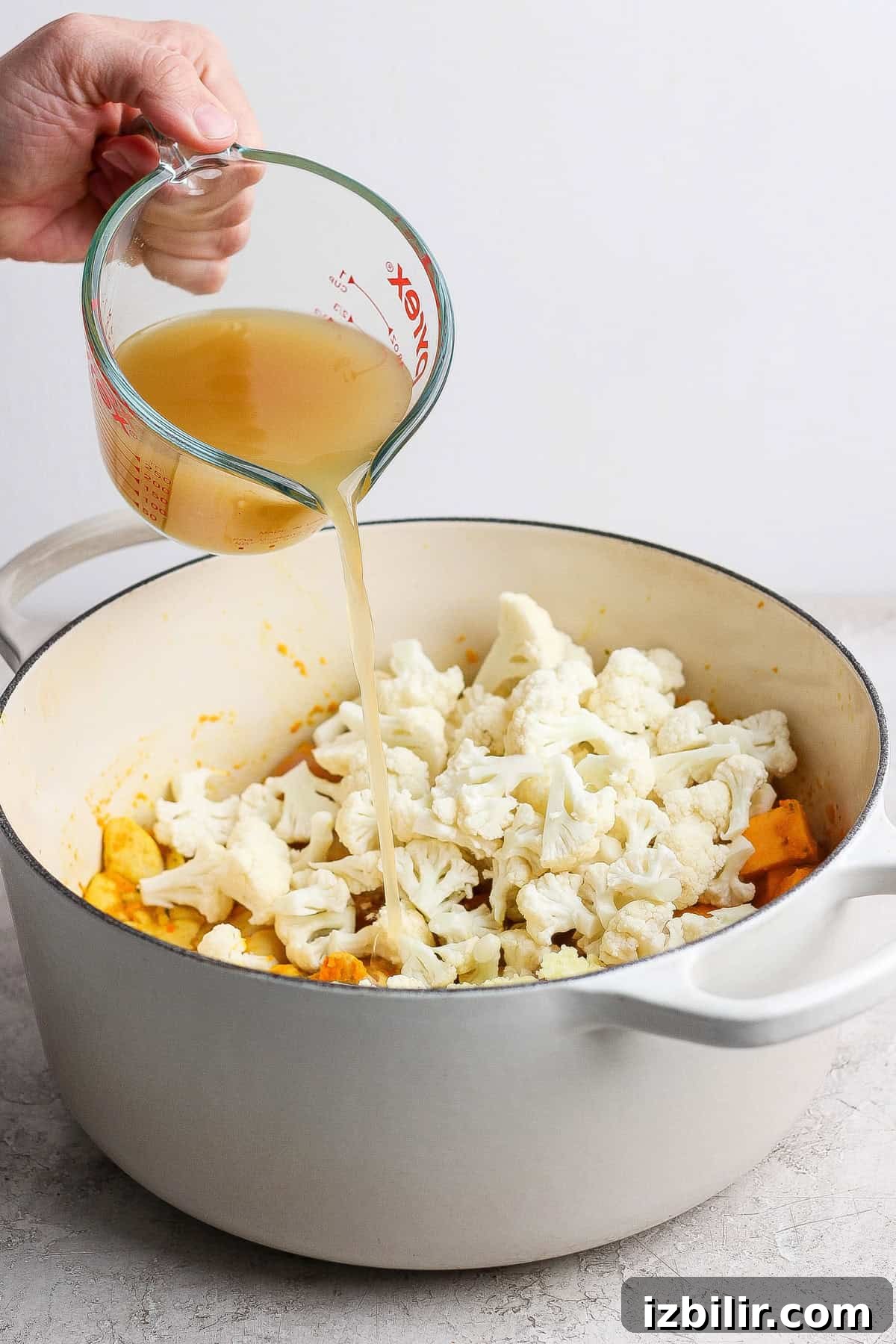 Comforting Soup to Boost Immunity 6 A hand pours broth from a glass measuring cup into a white pot filled with cauliflower florets and other vegetables.