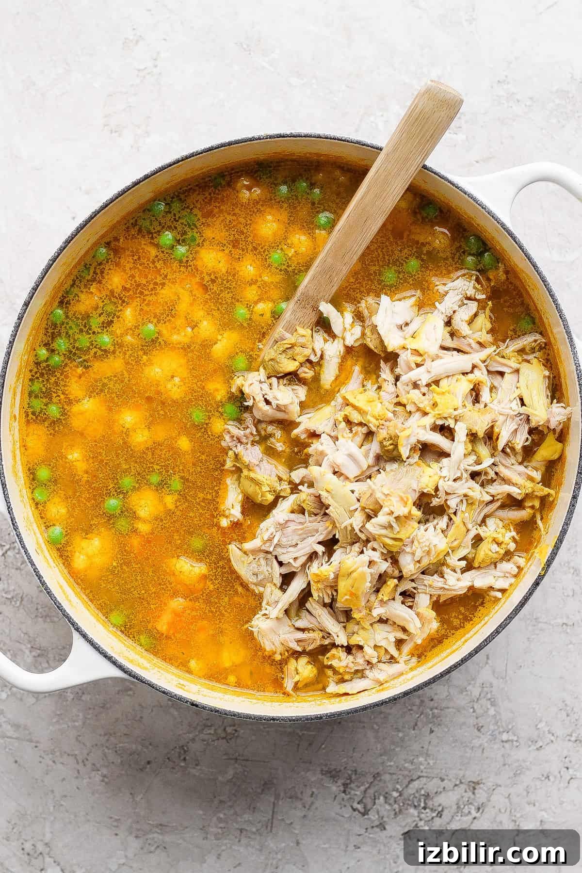 Comforting Soup to Boost Immunity 7 A pot of soup with shredded chicken, green peas, and a wooden spoon on a light-colored surface.