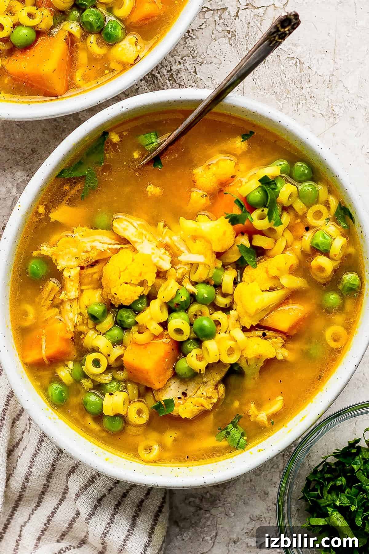 Comforting Soup to Boost Immunity 8 A bowl of chicken noodle soup with round pasta, peas, cauliflower, carrots, and herbs, with a spoon and a striped cloth nearby.