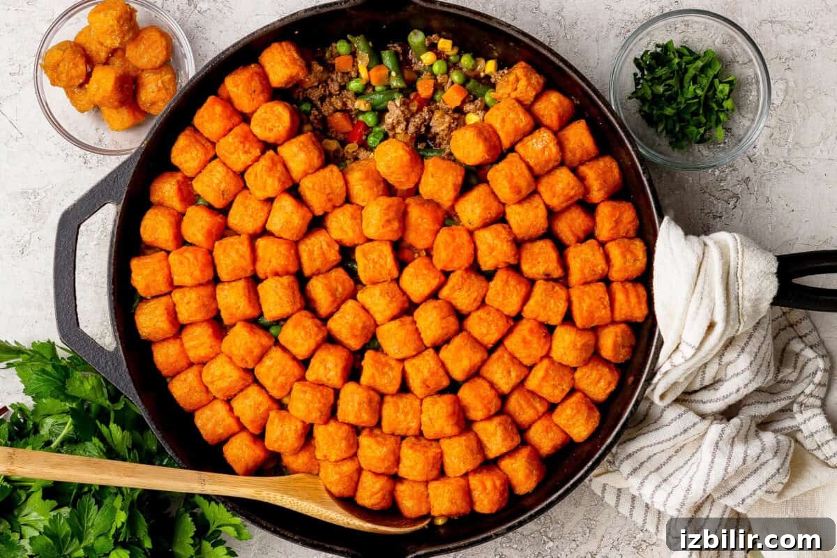 A skillet filled with tater tots arranged on top of a mixture of ground beef and vegetables, with bowls of parsley and extra tater tots nearby.