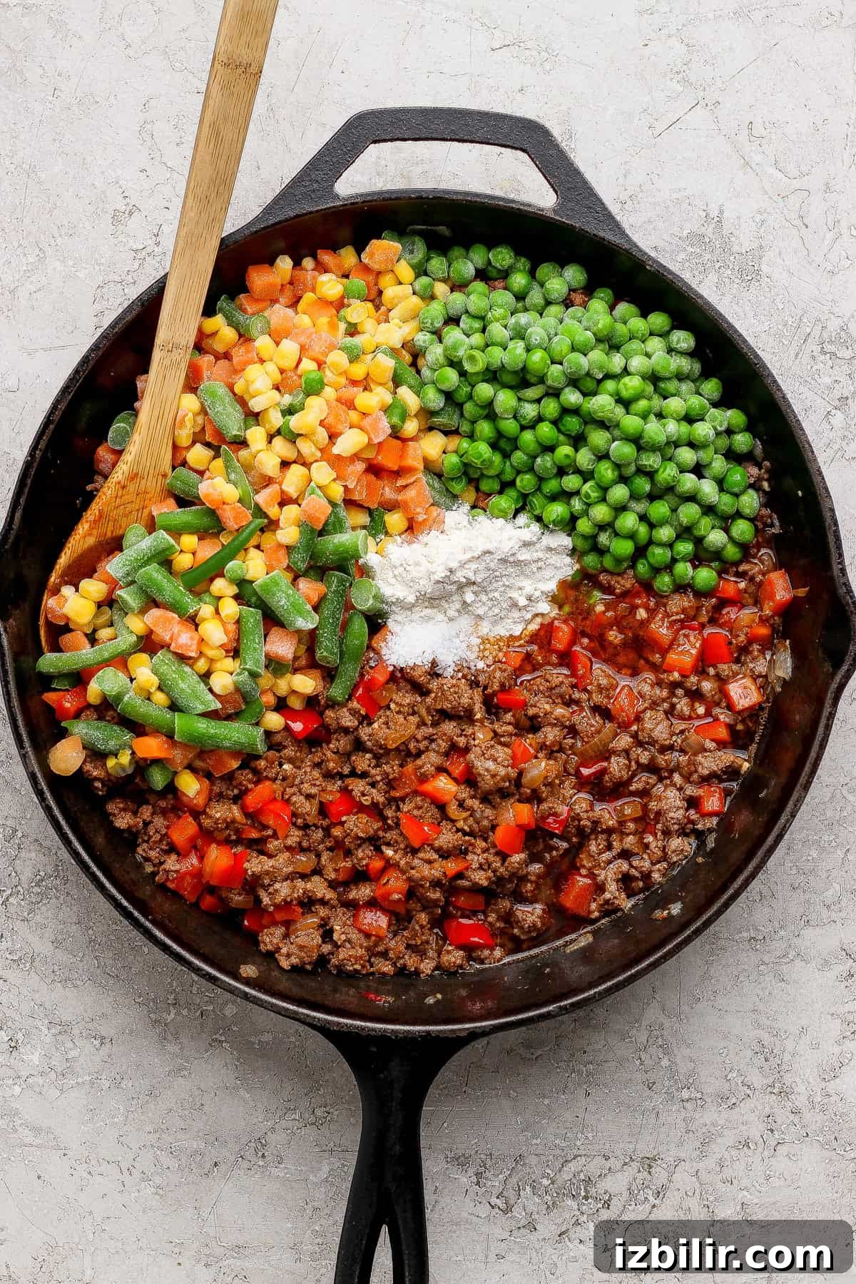 A cast iron skillet containing ground beef, peas, corn, green beans, diced red peppers, flour, and sauce, with a wooden spoon resting inside.