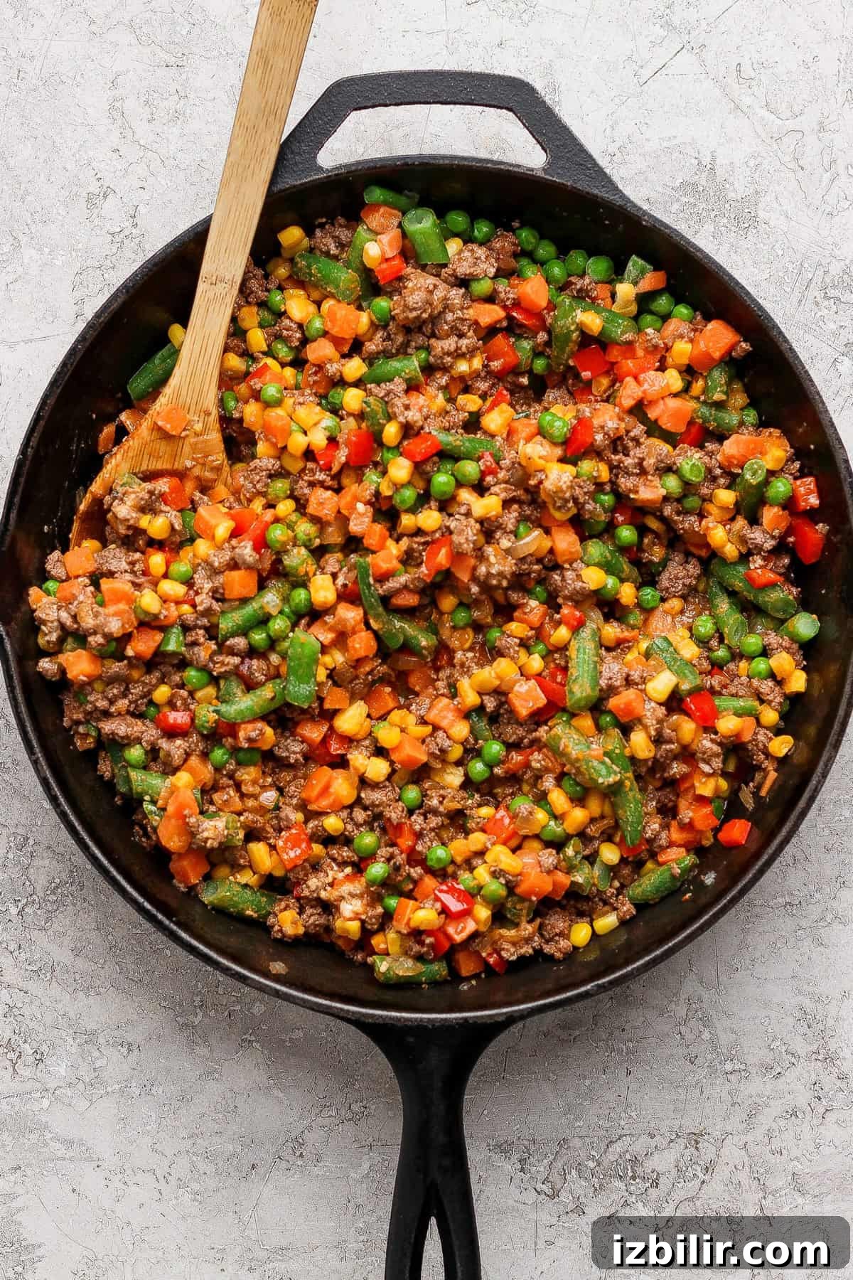 A cast iron skillet filled with cooked ground beef mixed with assorted vegetables, including peas, corn, carrots, and green beans, with a wooden spatula.