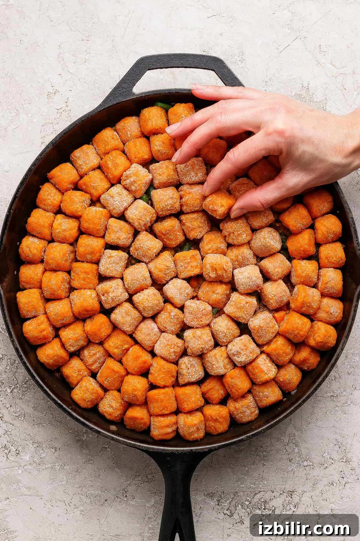 A hand arranges frozen tater tots in a single layer inside a round cast iron skillet on a light textured surface.