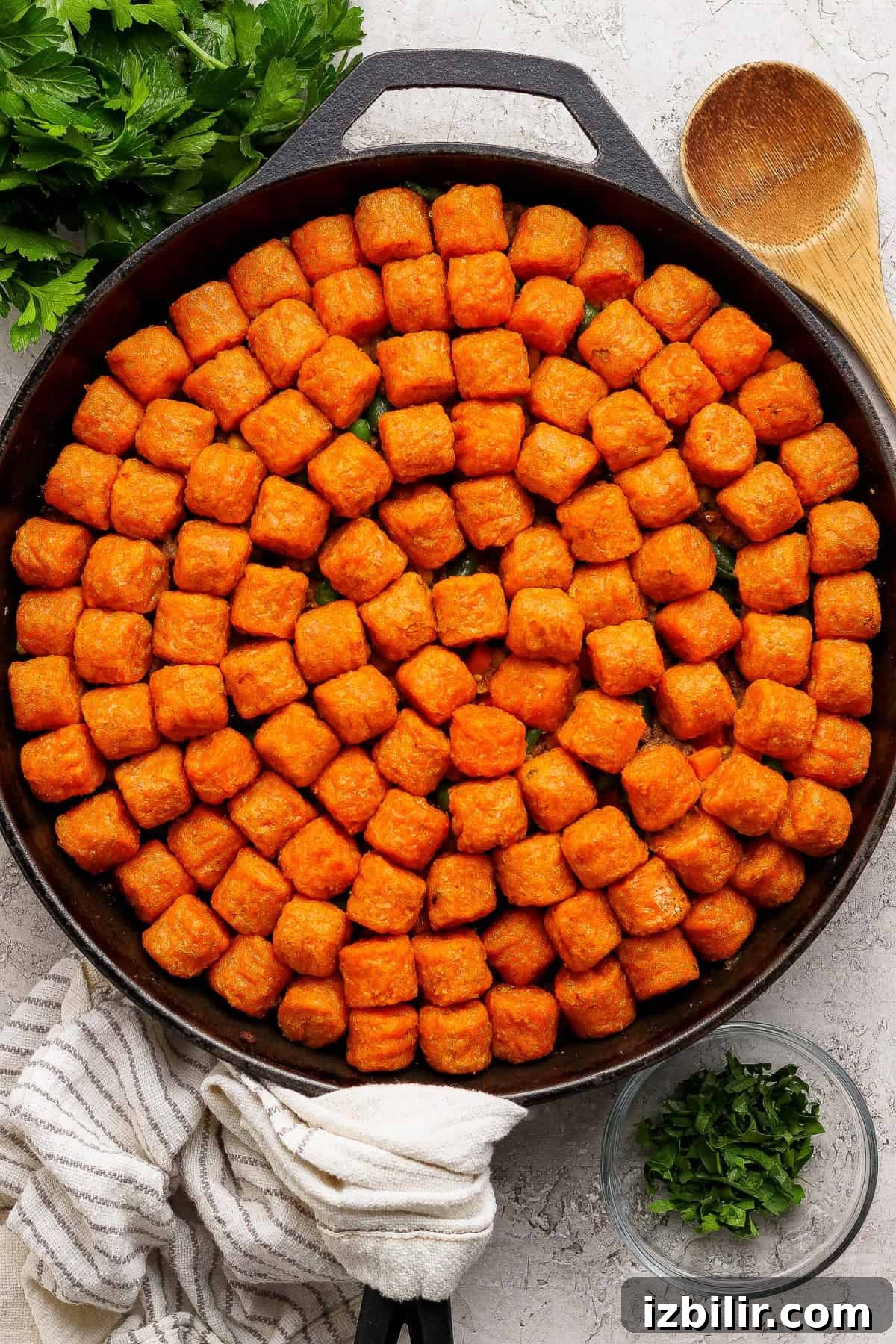 A cast iron skillet filled with evenly arranged tater tots, surrounded by a wooden spoon, parsley, and a striped kitchen towel.