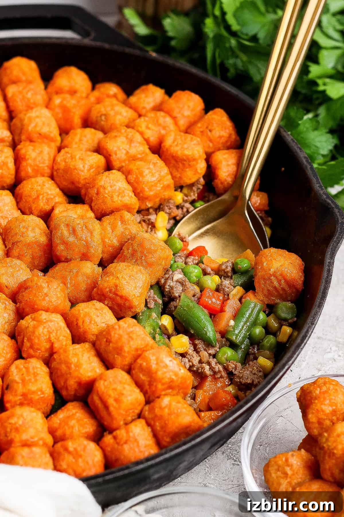 A cast iron skillet filled with ground beef, mixed vegetables, and topped with rows of tater tots. A gold serving spoon is in the skillet.