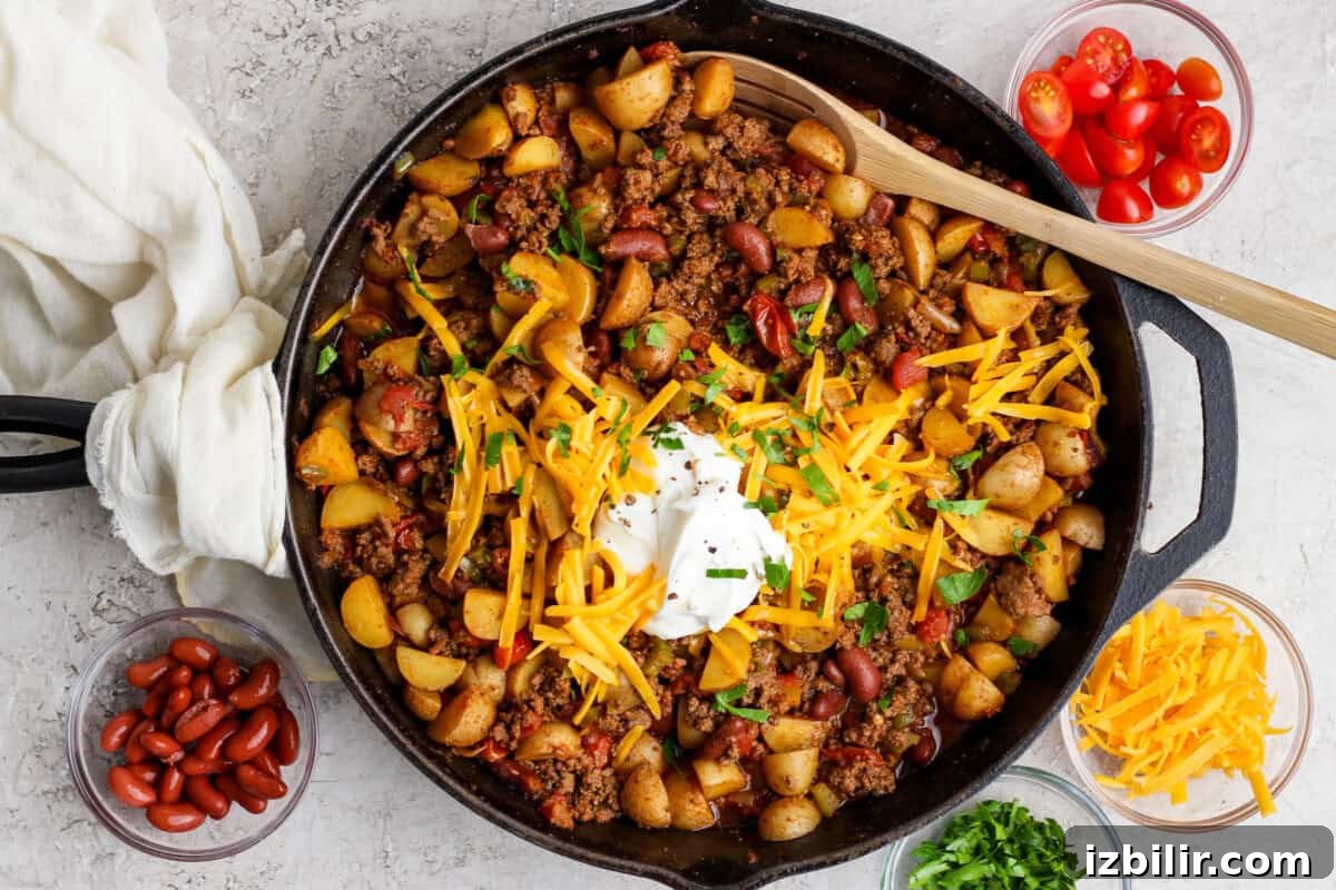 A cast iron skillet filled with chili, potatoes, kidney beans, ground meat, topped with shredded cheese, sour cream, and herbs. Bowls of tomatoes, beans, cheese, and herbs surround the skillet.