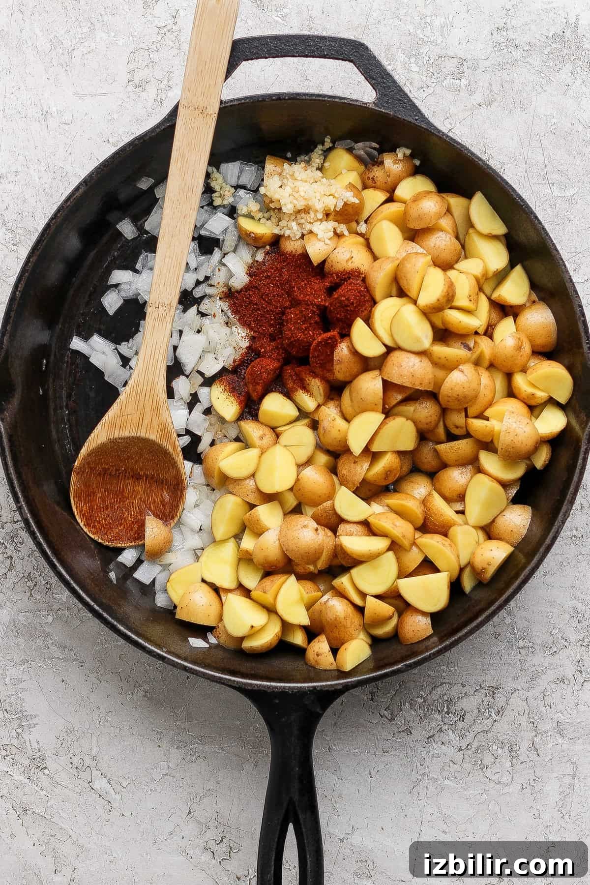 A cast iron skillet filled with chopped potatoes, diced onions, minced garlic, and paprika, with a wooden spoon resting on the side.