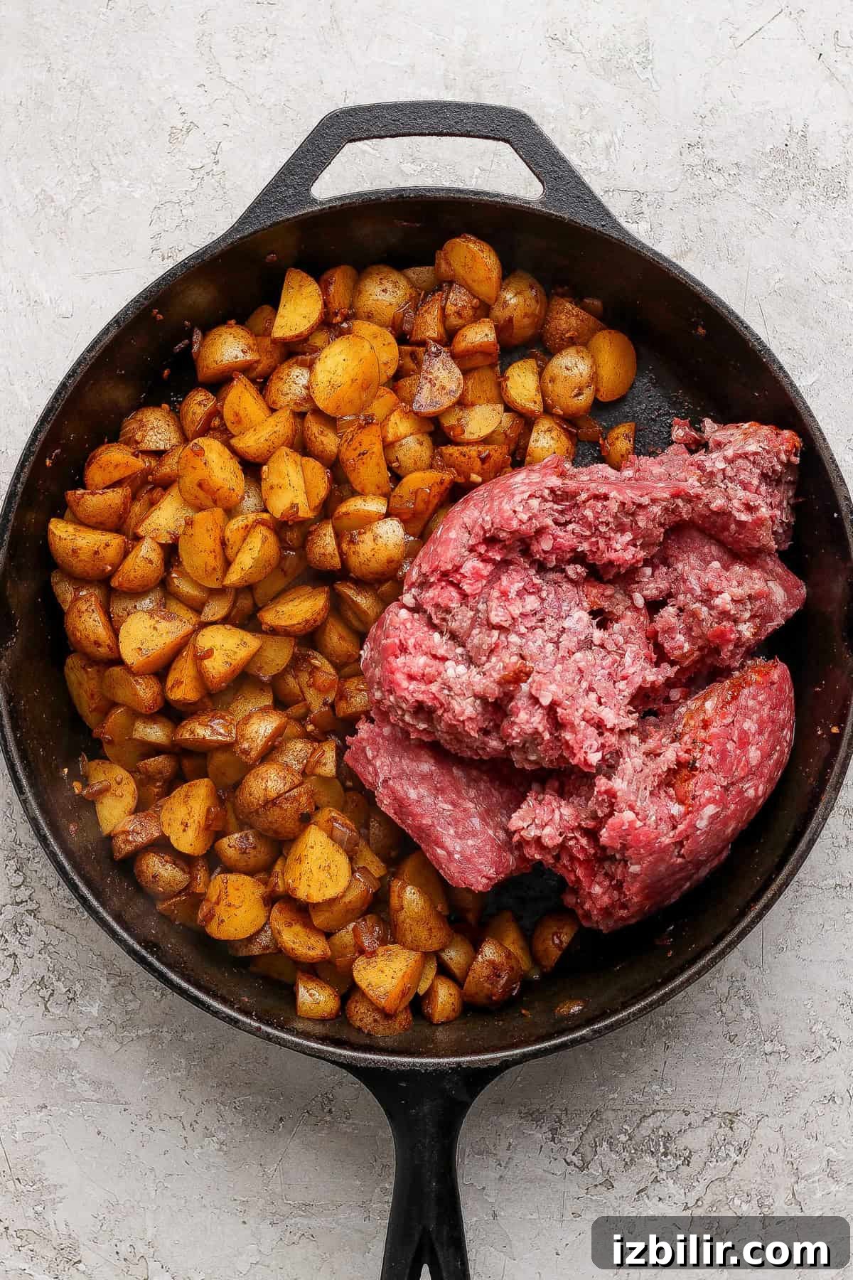 A cast iron skillet with cooked diced potatoes on one side and raw ground beef on the other, on a light textured surface.