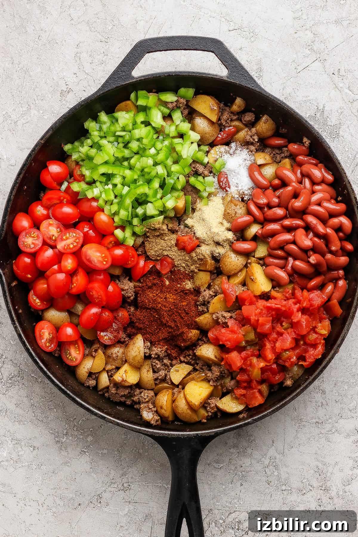 A cast iron skillet filled with diced potatoes, chopped tomatoes, kidney beans, green bell peppers, grape tomatoes, spices, and ground meat, all arranged in sections before mixing.