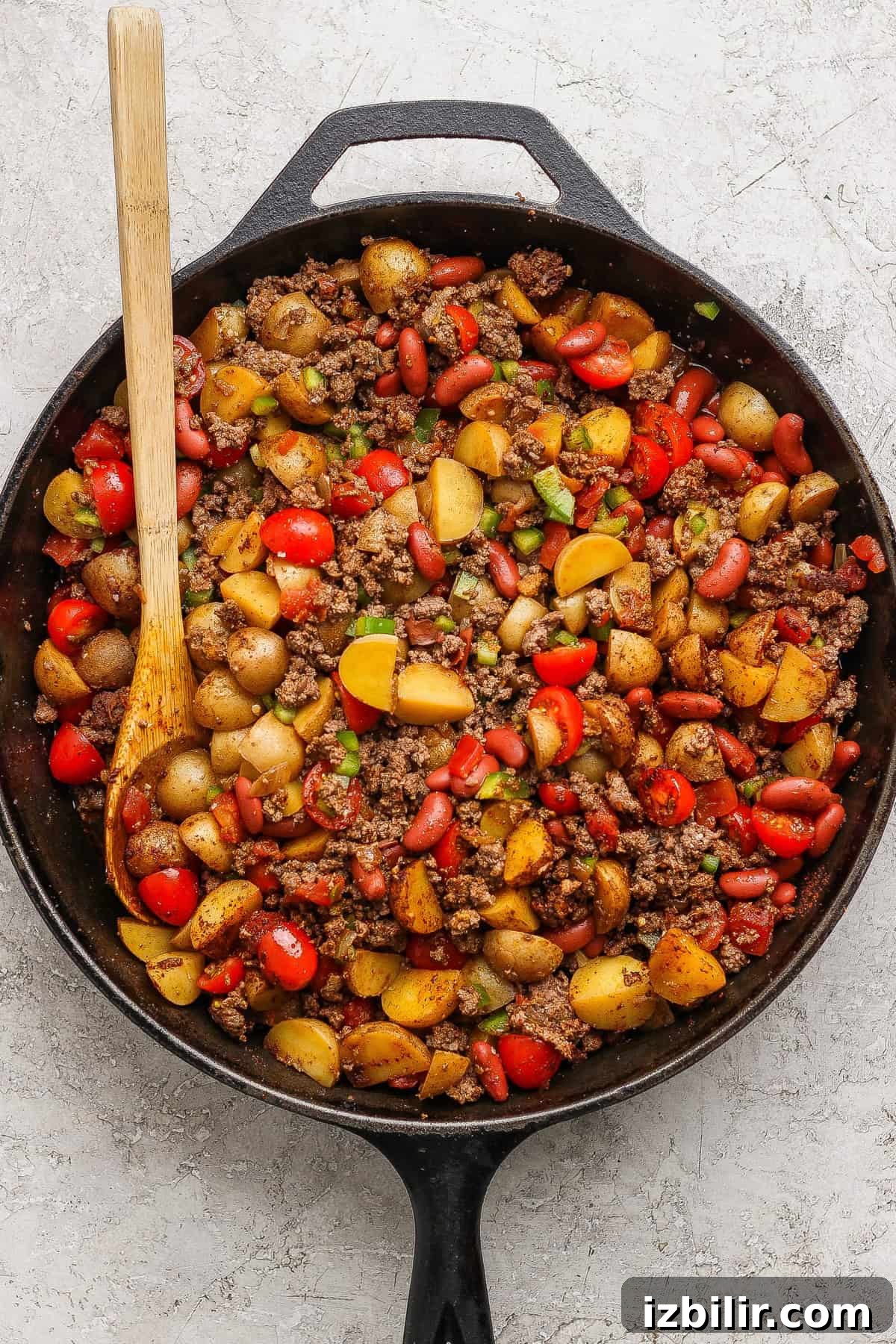 A cast iron skillet filled with cooked ground beef, halved baby potatoes, kidney beans, cherry tomatoes, and chopped green onions, with a wooden spoon resting inside.