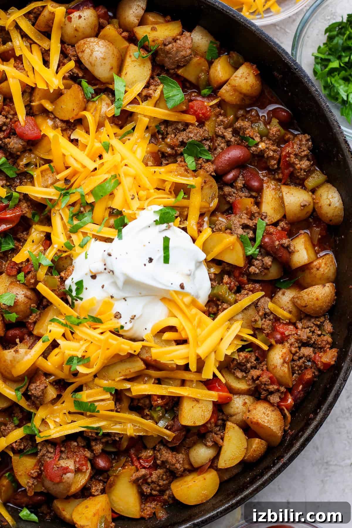 A skillet filled with ground beef, kidney beans, potatoes, and tomatoes, topped with shredded cheddar cheese, sour cream, and chopped parsley.
