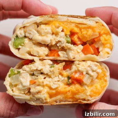 Two hands hold a halved breakfast burrito filled with scrambled eggs, cheese, onions, green peppers, and orange bell peppers.