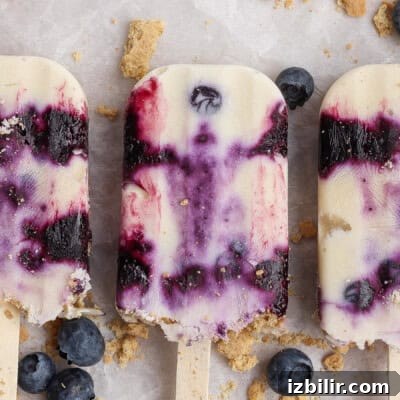 Three creamy blueberry popsicles with purple swirls are arranged on a light surface, surrounded by scattered blueberries and crumbled cookie pieces.