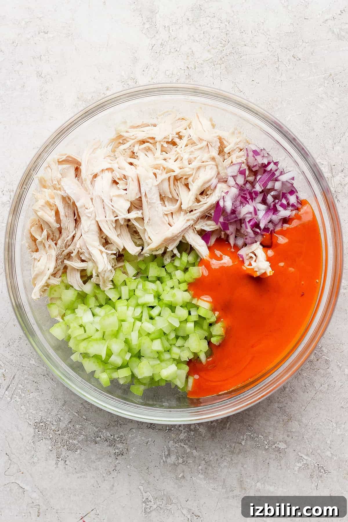A glass bowl containing shredded chicken, chopped celery, diced red onion, and orange buffalo sauce, arranged in separate sections before mixing.