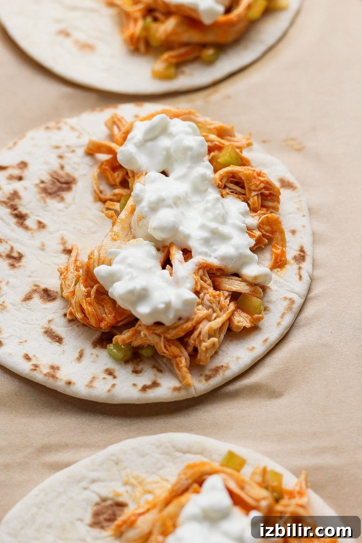 Three flour tortillas topped with shredded chicken, diced vegetables, and a dollop of white sauce, arranged on a light brown surface.