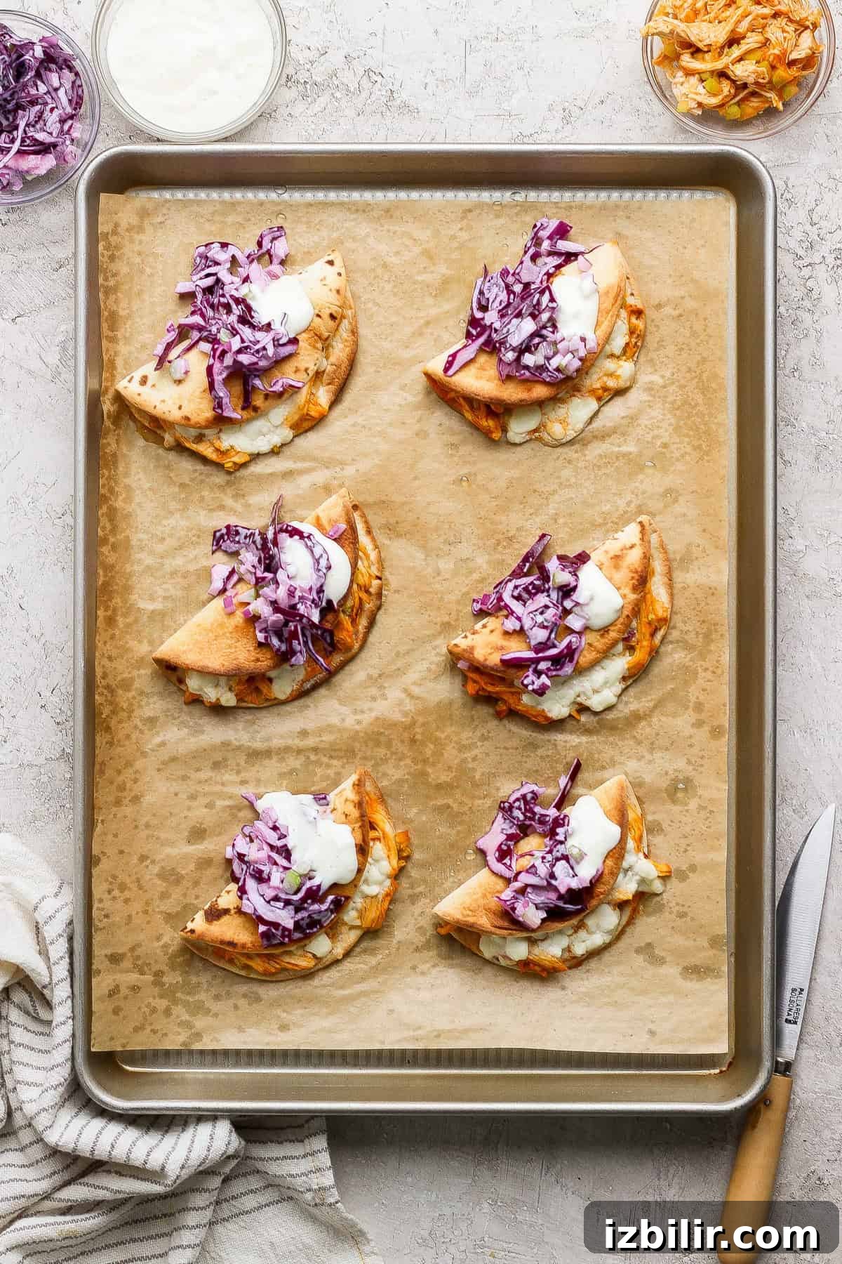Six folded pita tacos filled with shredded meat, purple cabbage slaw, and a dollop of white sauce arranged on a parchment-lined baking sheet.