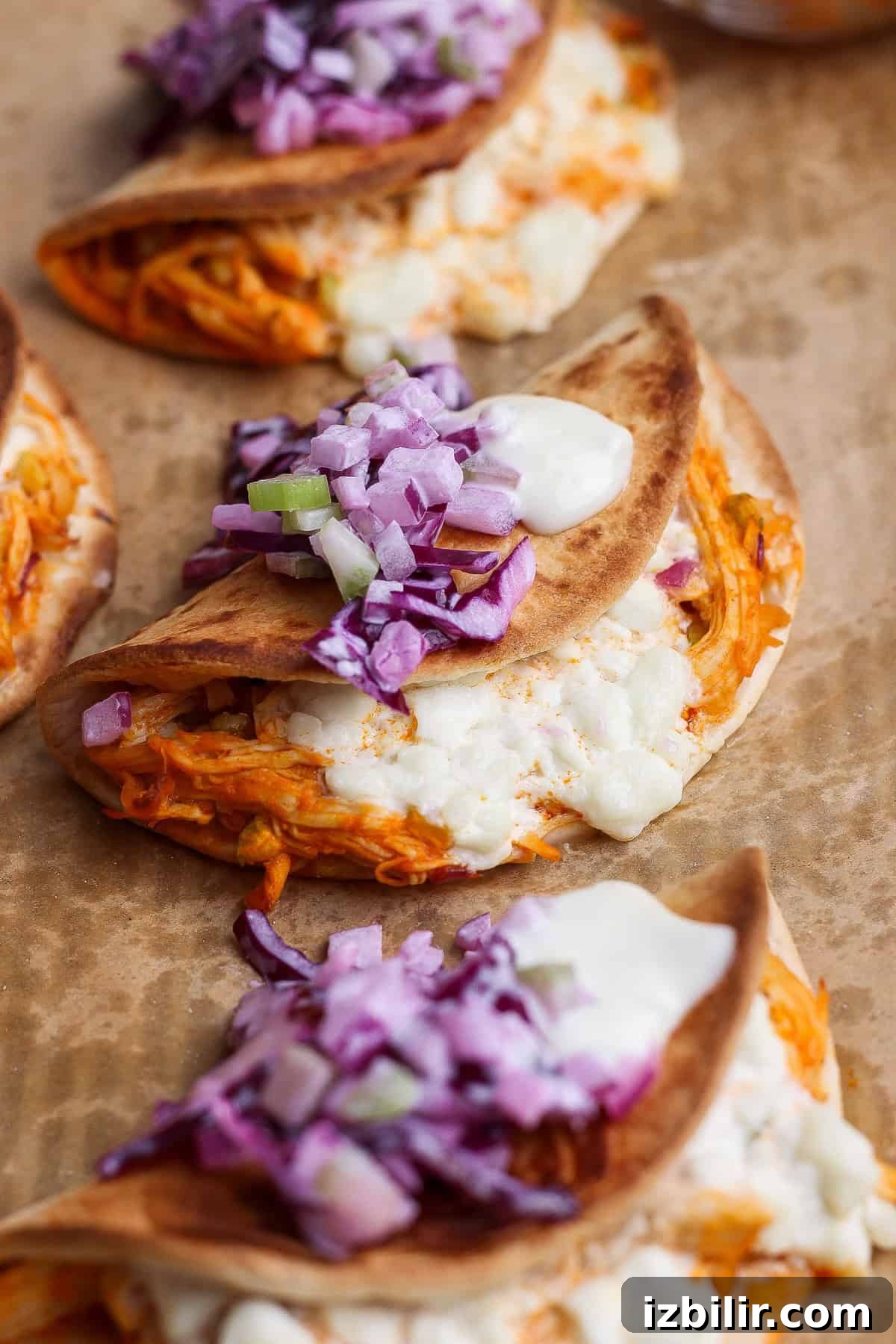 Three folded tacos filled with shredded meat and melted cheese, topped with chopped red onions and a dollop of white sauce, placed on brown parchment paper.