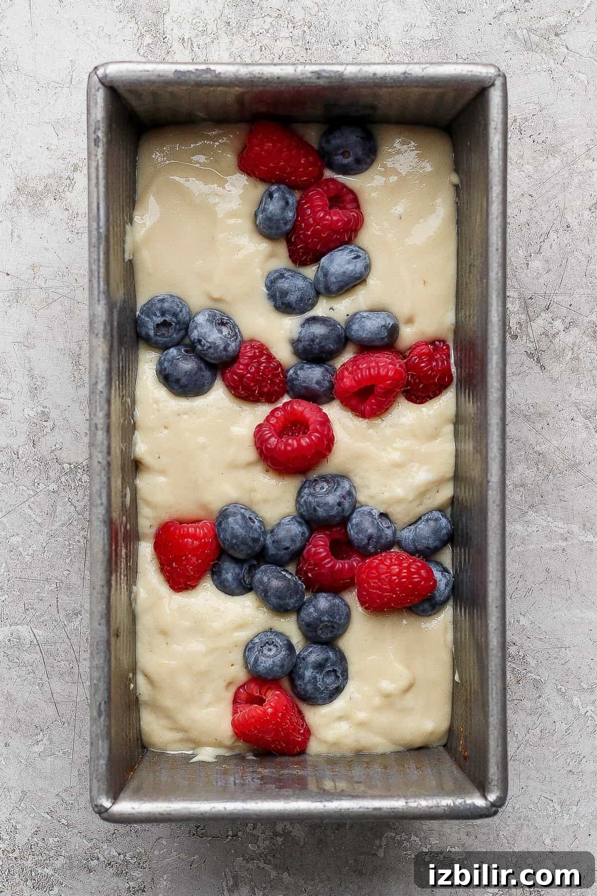 High-Protein Buns: Triple Berry and Raspberry Almond Croissant 5 A loaf pan filled with cake batter, topped with scattered fresh blueberries and raspberries, ready to be baked.