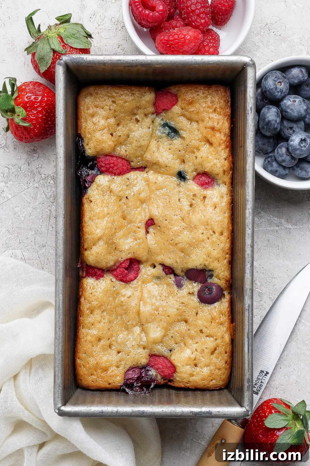 High-Protein Buns: Triple Berry and Raspberry Almond Croissant 6 A loaf of berry bread with visible raspberries and blueberries in a metal pan, surrounded by fresh berries, a white cloth, and a knife.
