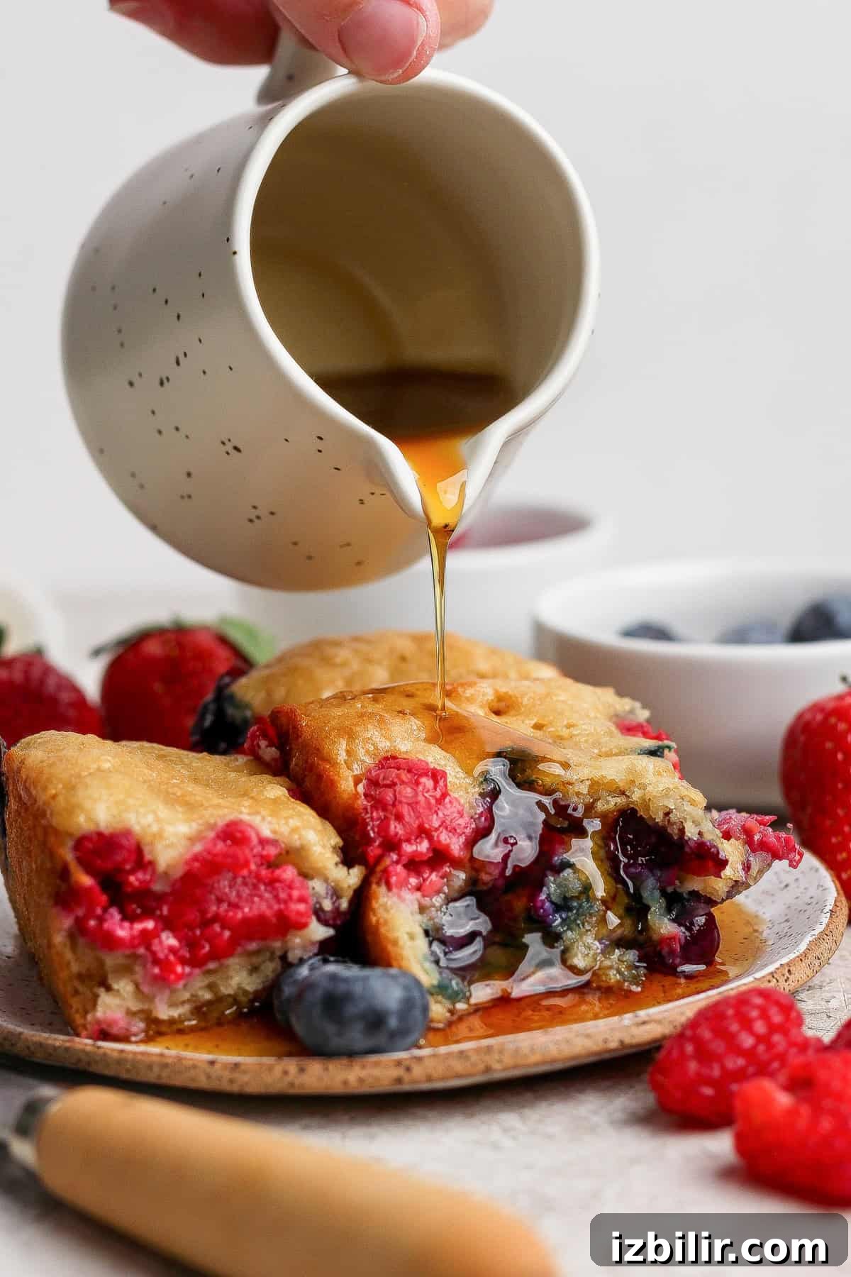 High-Protein Buns: Triple Berry and Raspberry Almond Croissant 7 A hand pours syrup from a small white jug onto a stack of berry-filled pancakes on a plate, surrounded by fresh strawberries, raspberries, and blueberries.