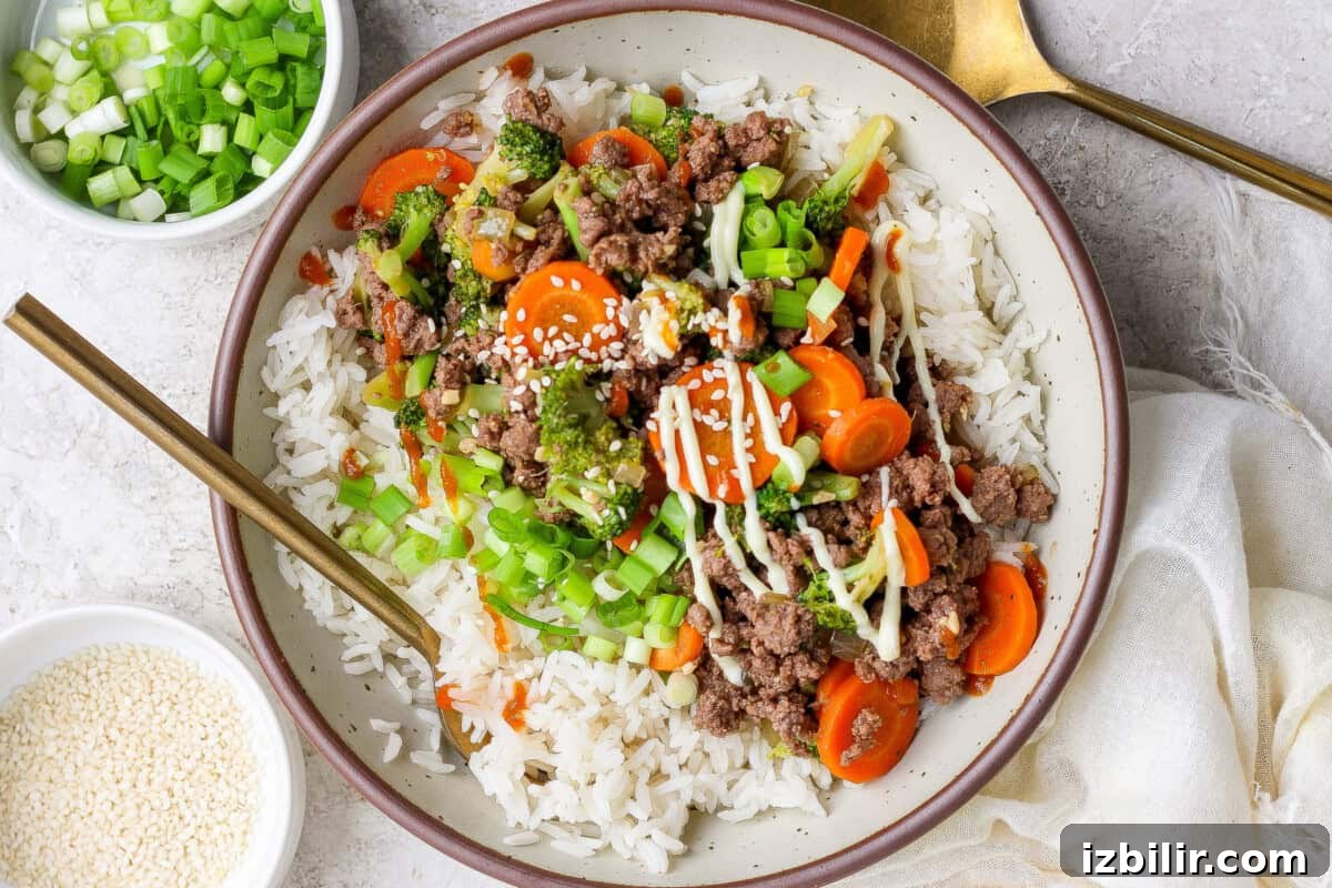 A bowl of white rice topped with ground beef, sliced carrots, broccoli, chopped green onions, sesame seeds, and a drizzle of sauce. Spoons and small bowls of green onions and sesame seeds nearby.