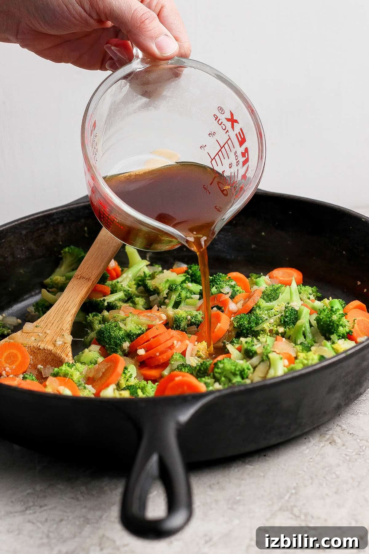 A hand pours sauce from a measuring cup into a cast iron skillet with broccoli and sliced carrots being stirred with a wooden spoon.