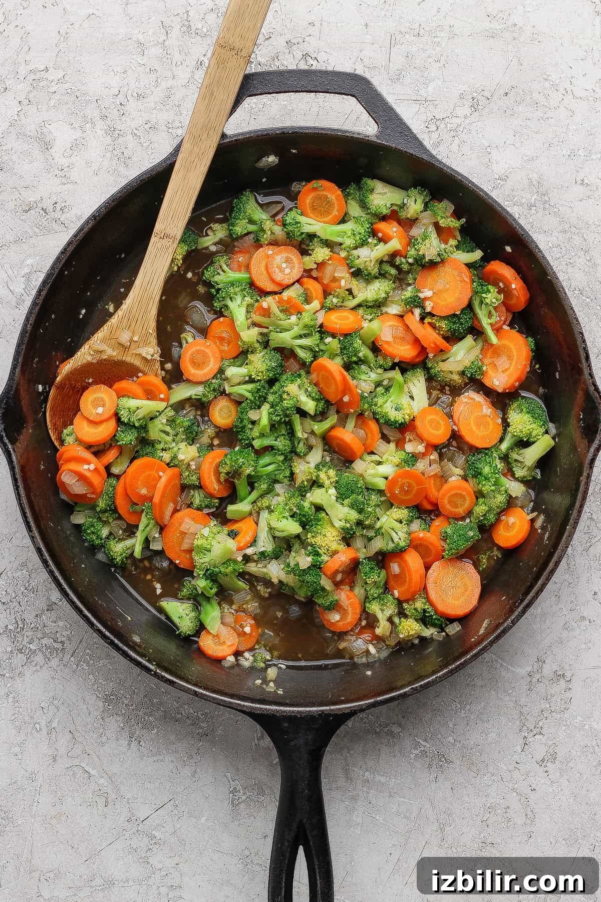 A cast iron skillet with sautéed broccoli and sliced carrots being stirred with a wooden spoon on a light textured surface.