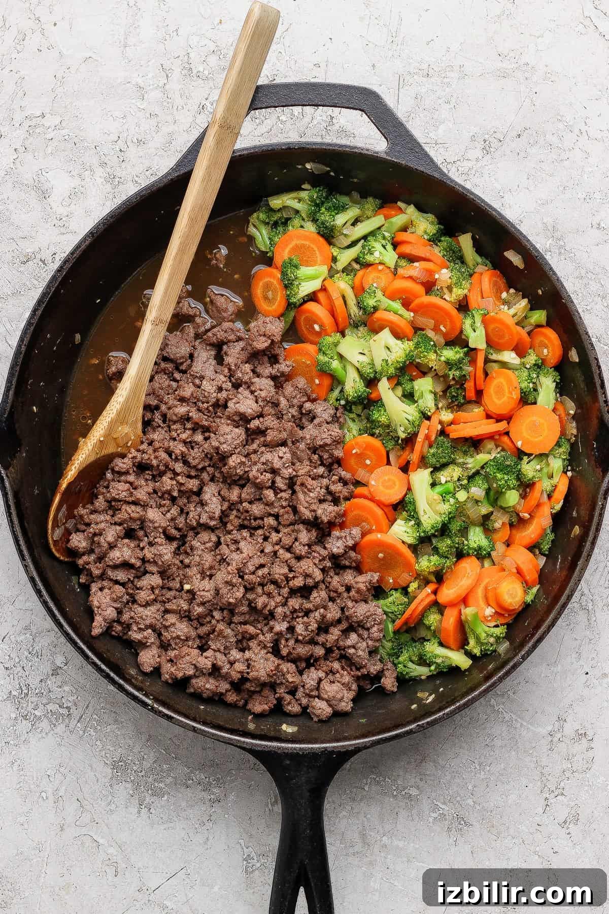A cast iron skillet contains cooked ground beef and sautéed broccoli and carrots, with a wooden spoon resting inside.