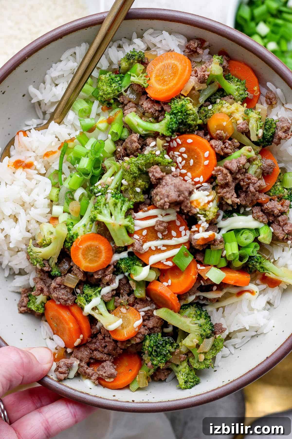A bowl of white rice topped with ground beef, sliced carrots, broccoli, green onions, and sesame seeds, with chopsticks resting on the edge.