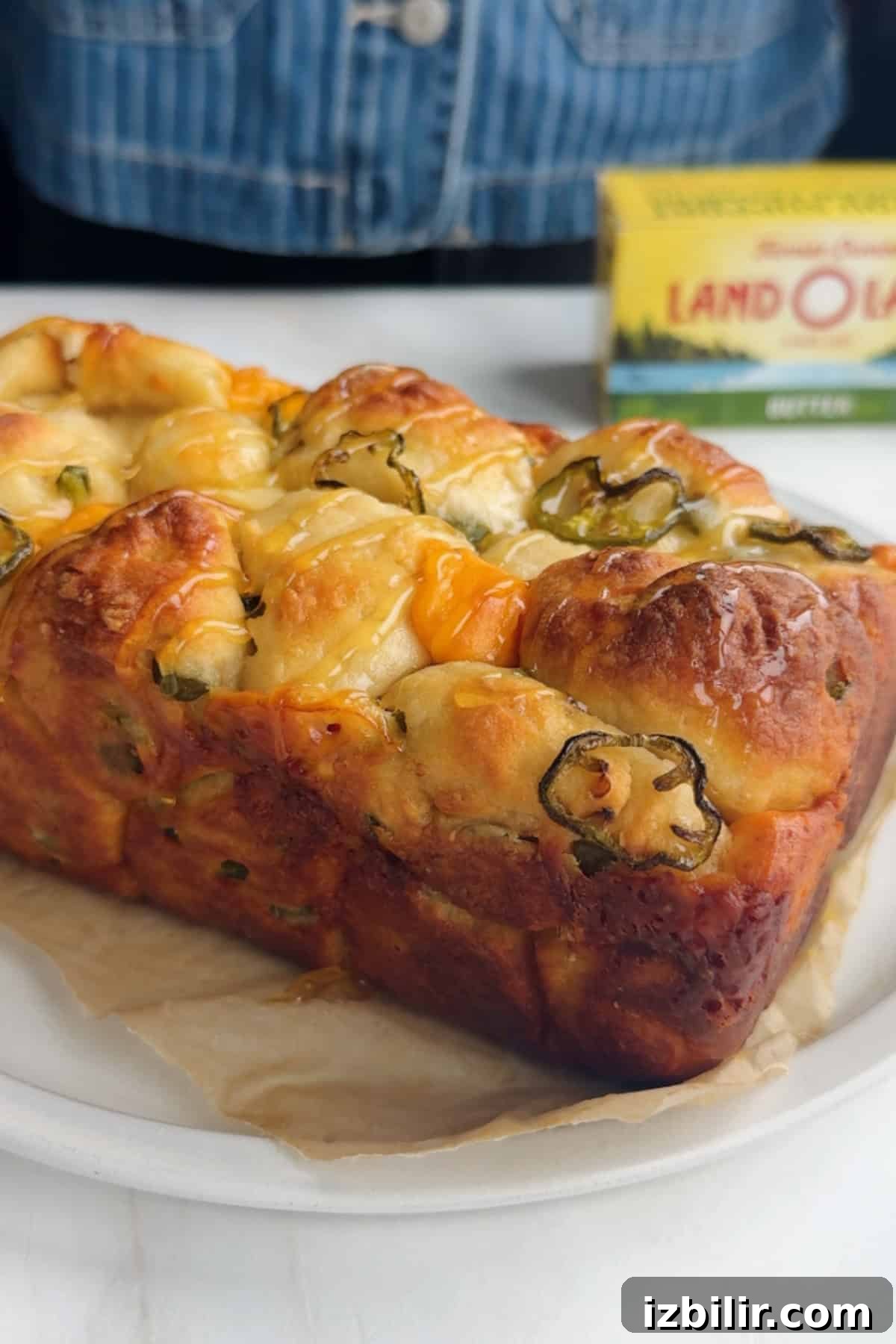 A loaf of savory bread with visible cheese and jalapeño slices rests on parchment paper, with a box of butter in the background.