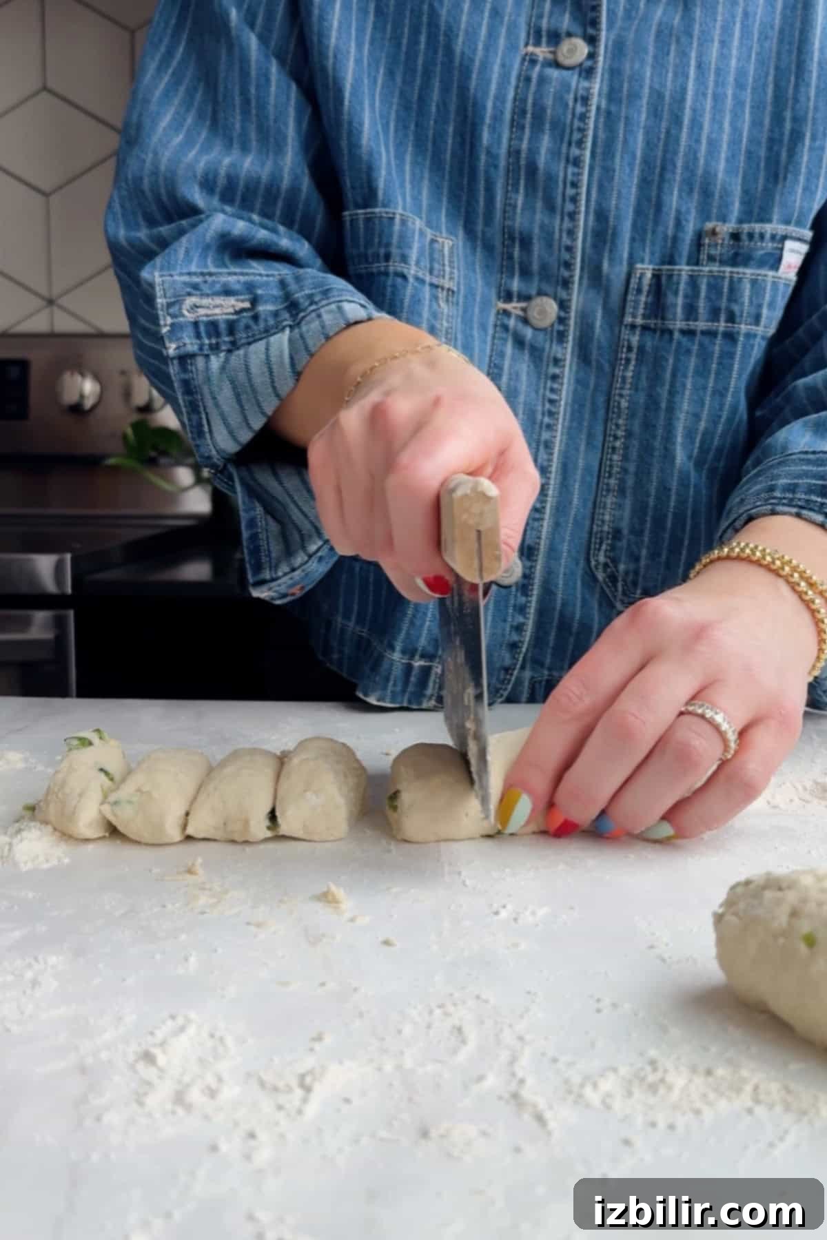 A person in a denim shirt cuts a roll of dough into pieces on a floured surface in a kitchen.