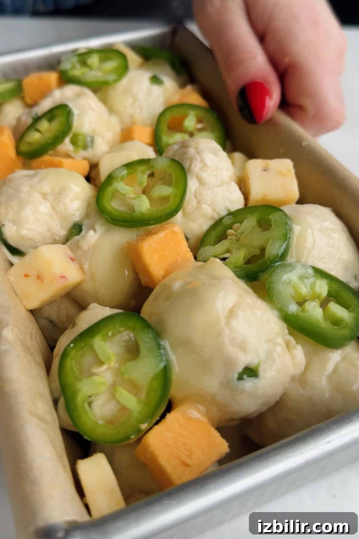 Close-up of unbaked dough balls in a pan, topped with sliced jalapeños and cubes of cheddar and pepper jack cheese.