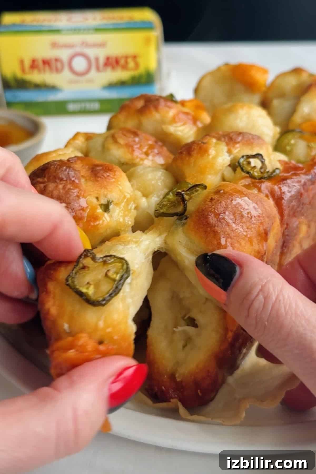 Two hands pull apart a piece of baked bread topped with sliced jalapeños, with a package of butter visible in the background.