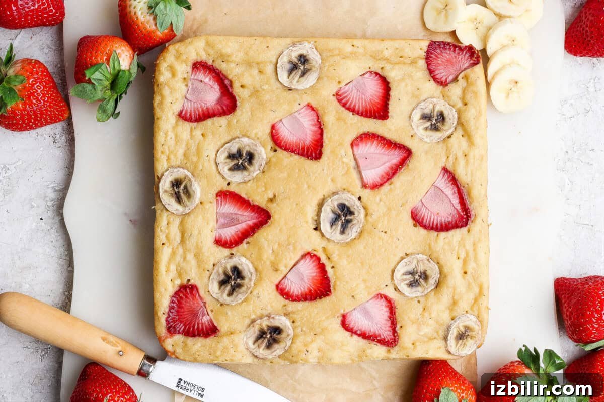 A square baked banana bread topped with sliced strawberries and bananas, surrounded by fresh strawberries and a knife on parchment paper.