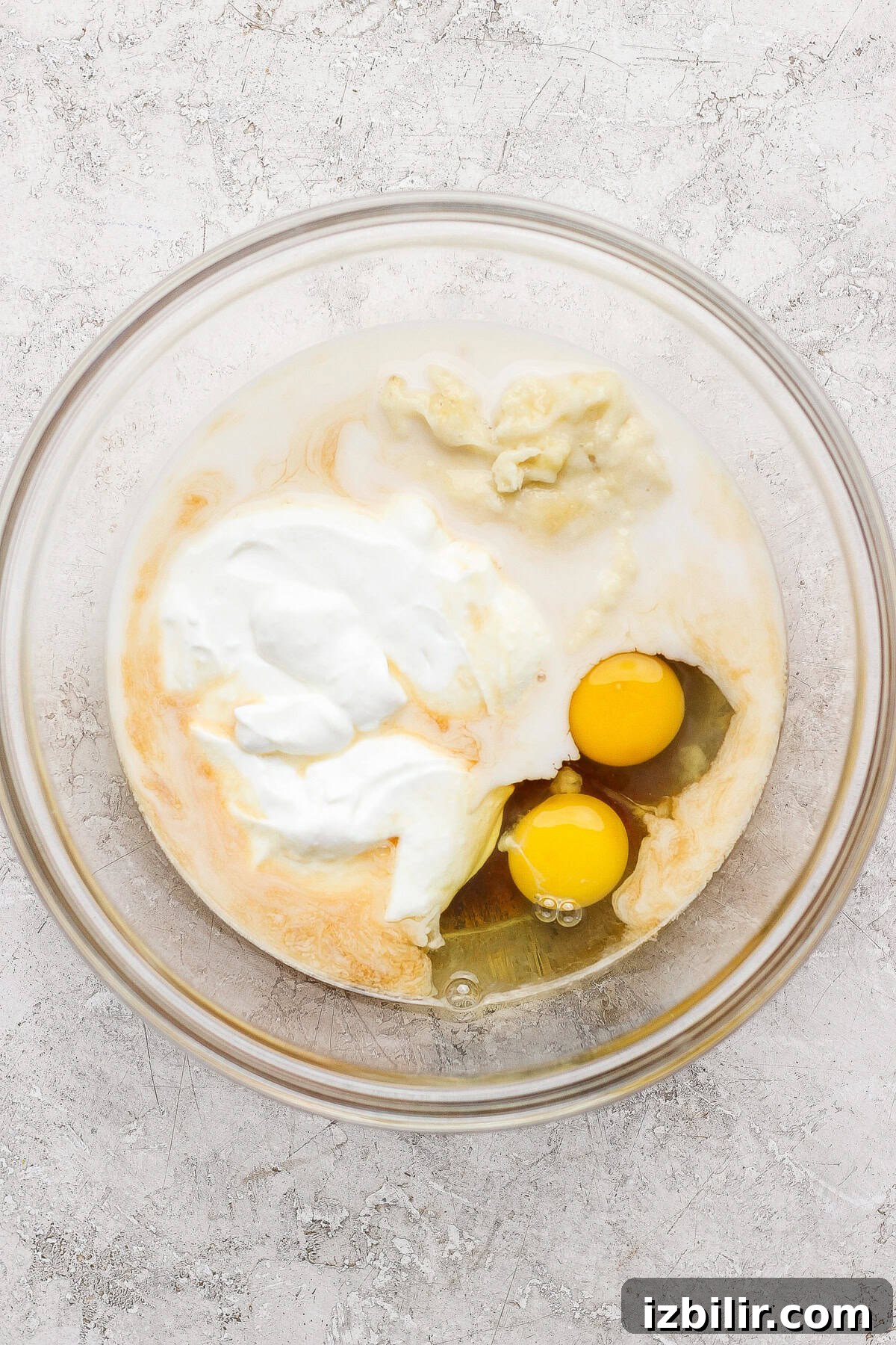 A glass bowl containing two raw eggs, yogurt, and other wet ingredients on a light textured surface.