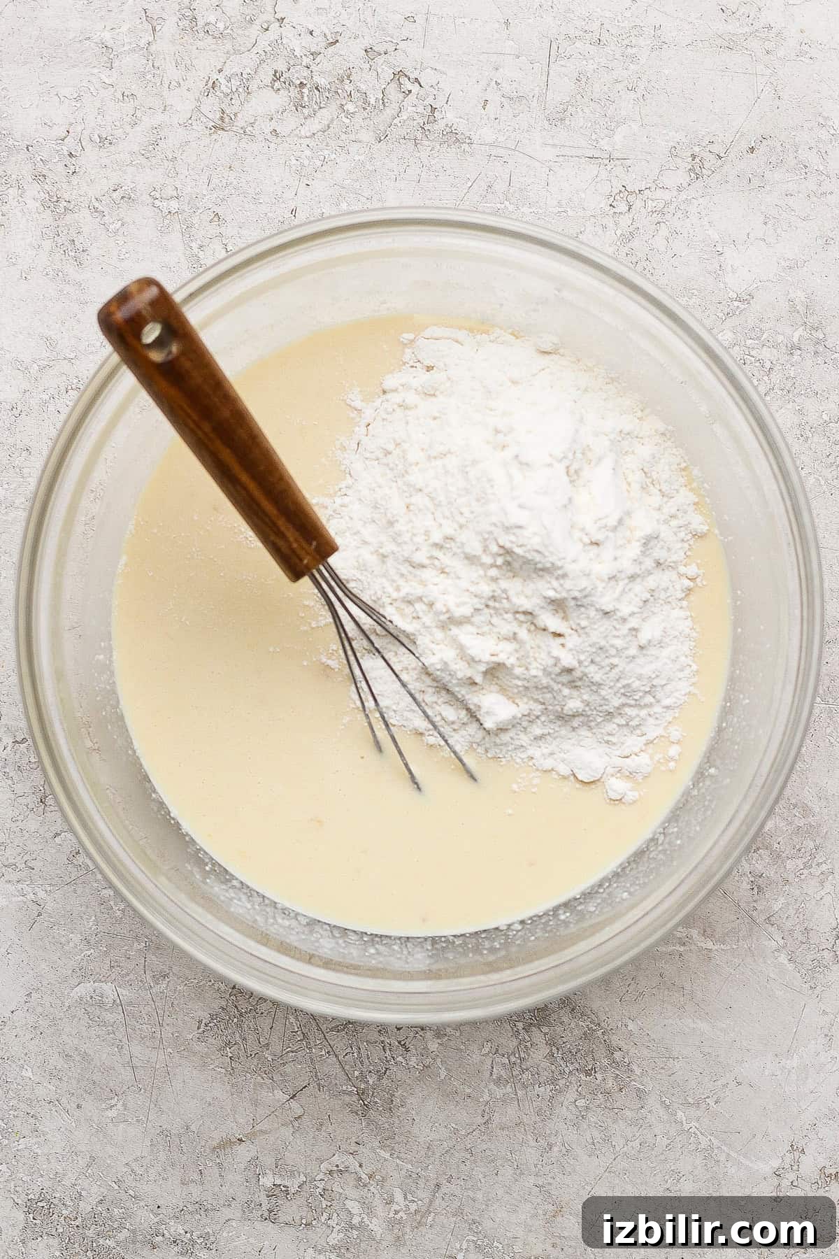 A glass bowl with liquid batter and a mound of flour, being mixed with a metal whisk with a wooden handle, on a light textured surface.