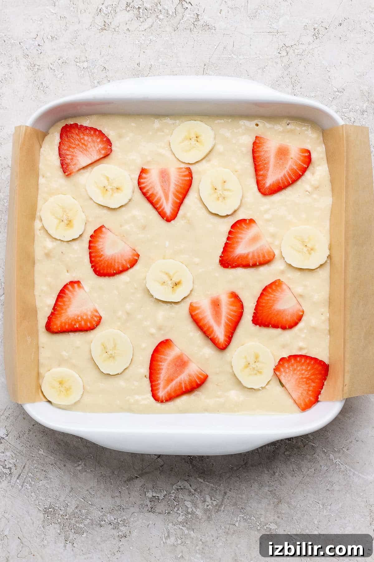 A baking dish lined with parchment paper contains unbaked batter topped with sliced strawberries and bananas.
