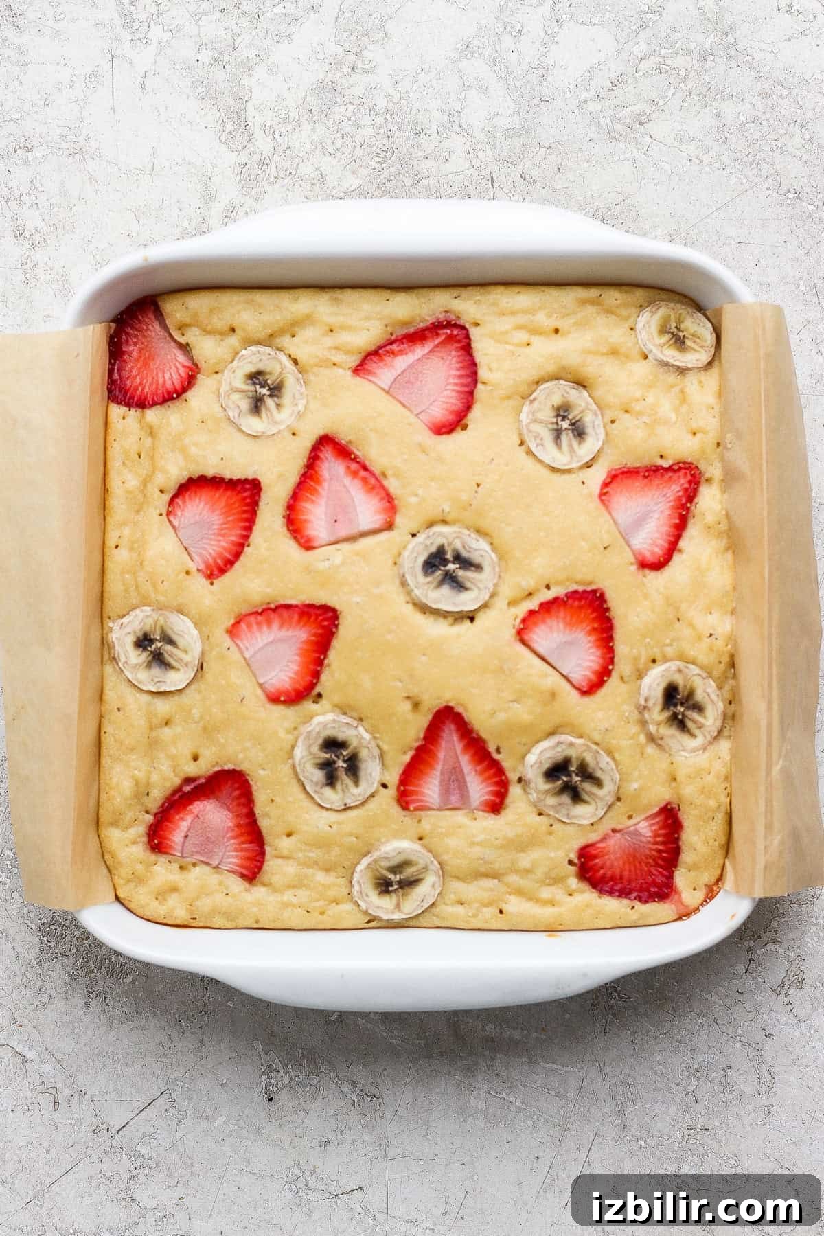 A square baking dish with a baked cake topped with sliced strawberries and bananas, lined with parchment paper on a light textured surface.