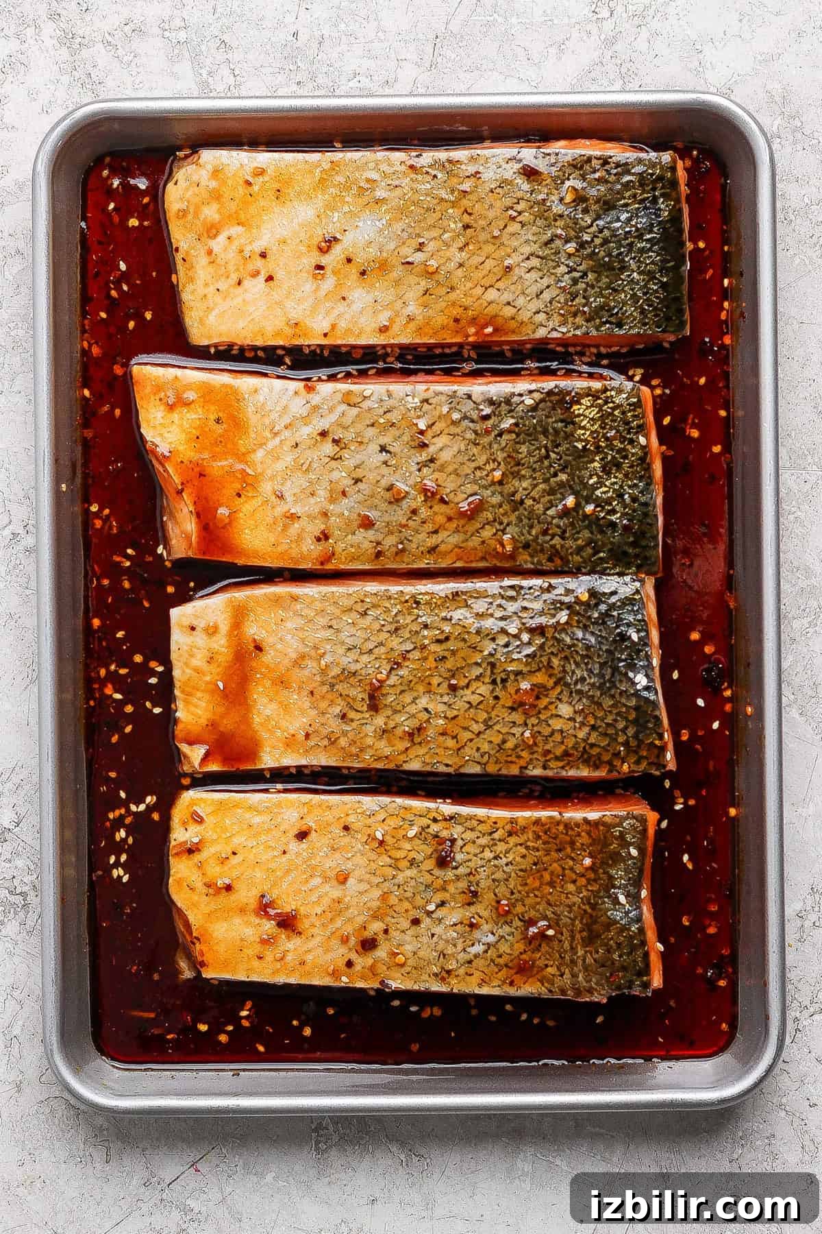 Four raw salmon fillets with skin on, arranged in a baking tray and marinated in a dark sauce with visible spices and chili flakes.
