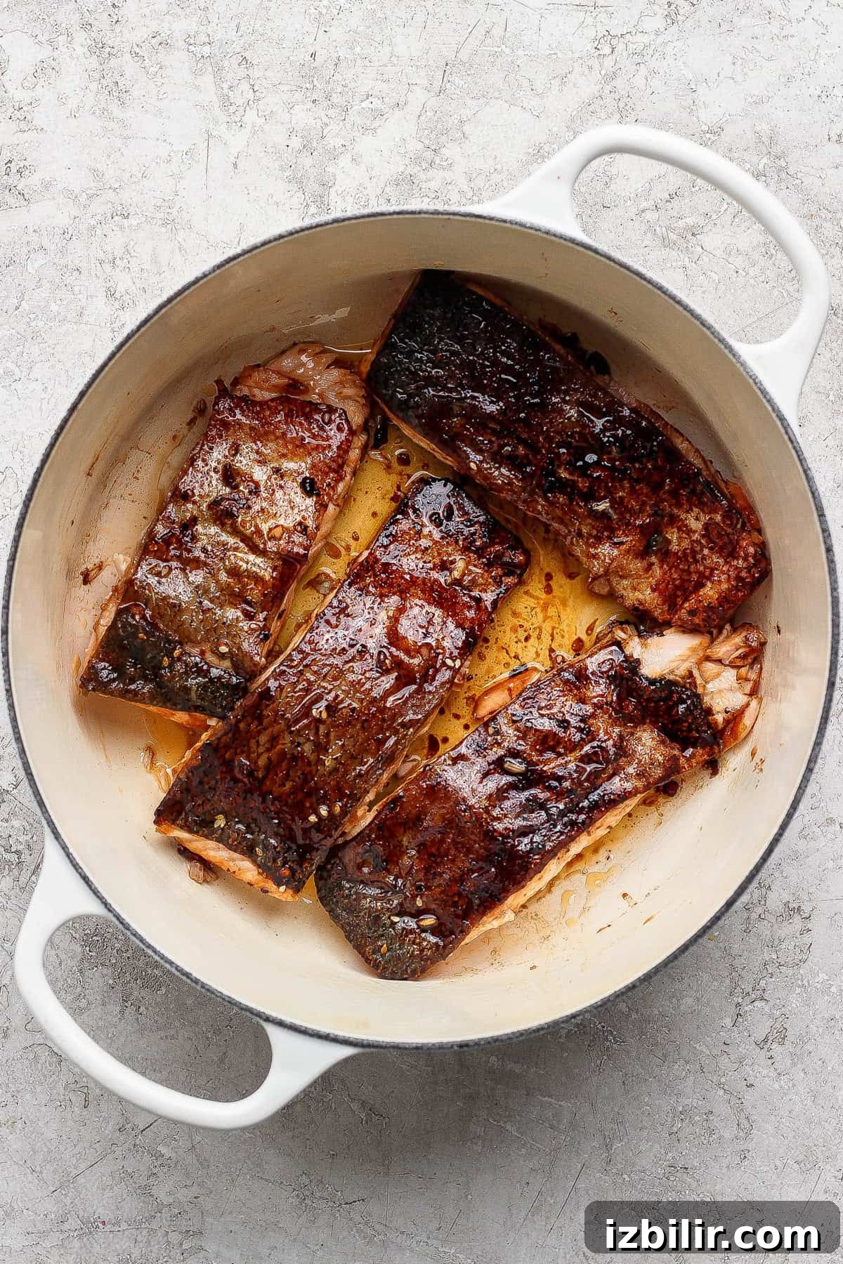 Four seared salmon fillets with browned skin cooking in a white enameled cast iron pan on a light textured surface.