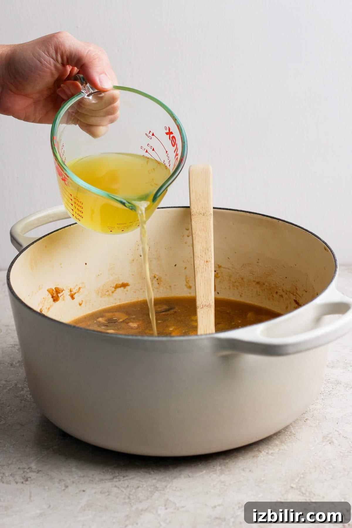 A hand pours broth from a measuring cup into a large pot containing a brown mixture, with a wooden spatula resting inside the pot.