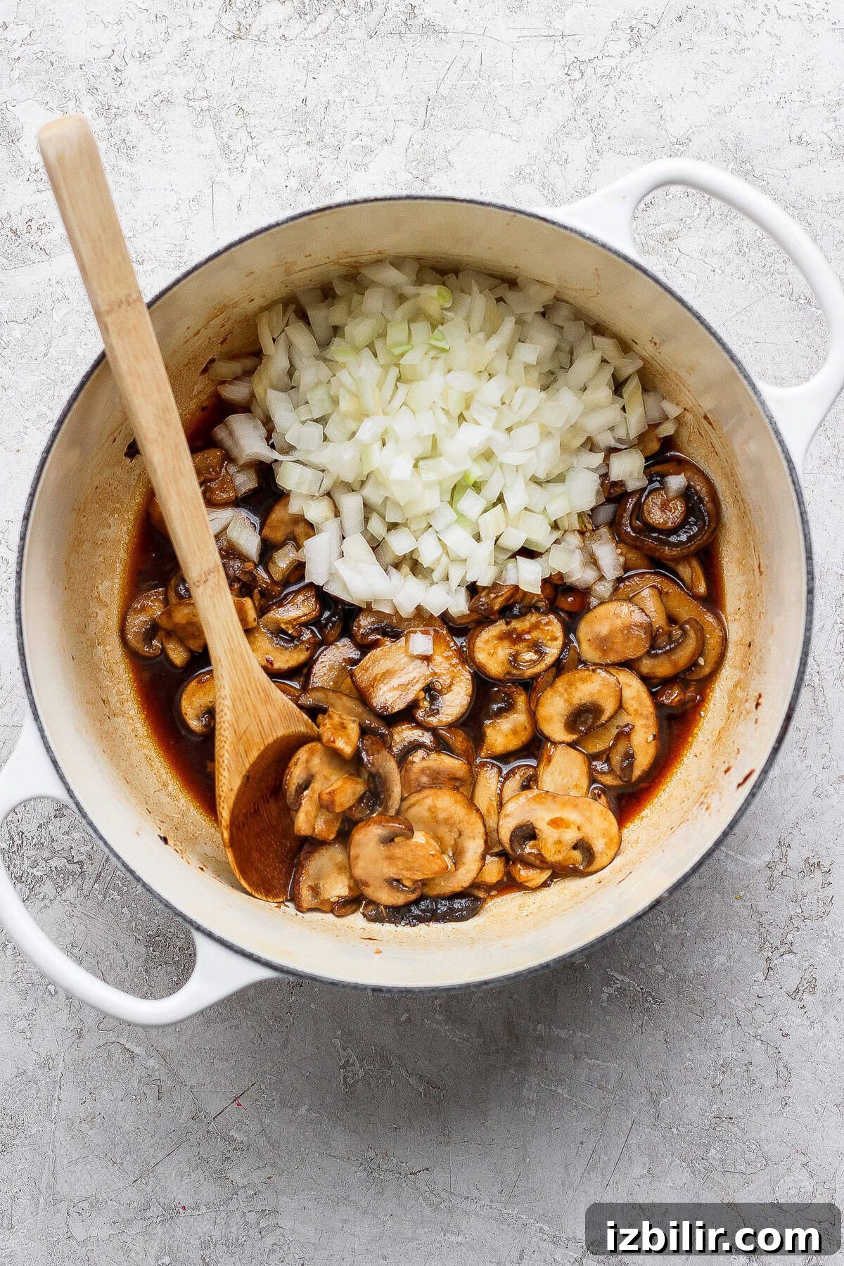 A white pot containing sliced mushrooms in sauce and a pile of diced onions, with a wooden spoon resting inside.