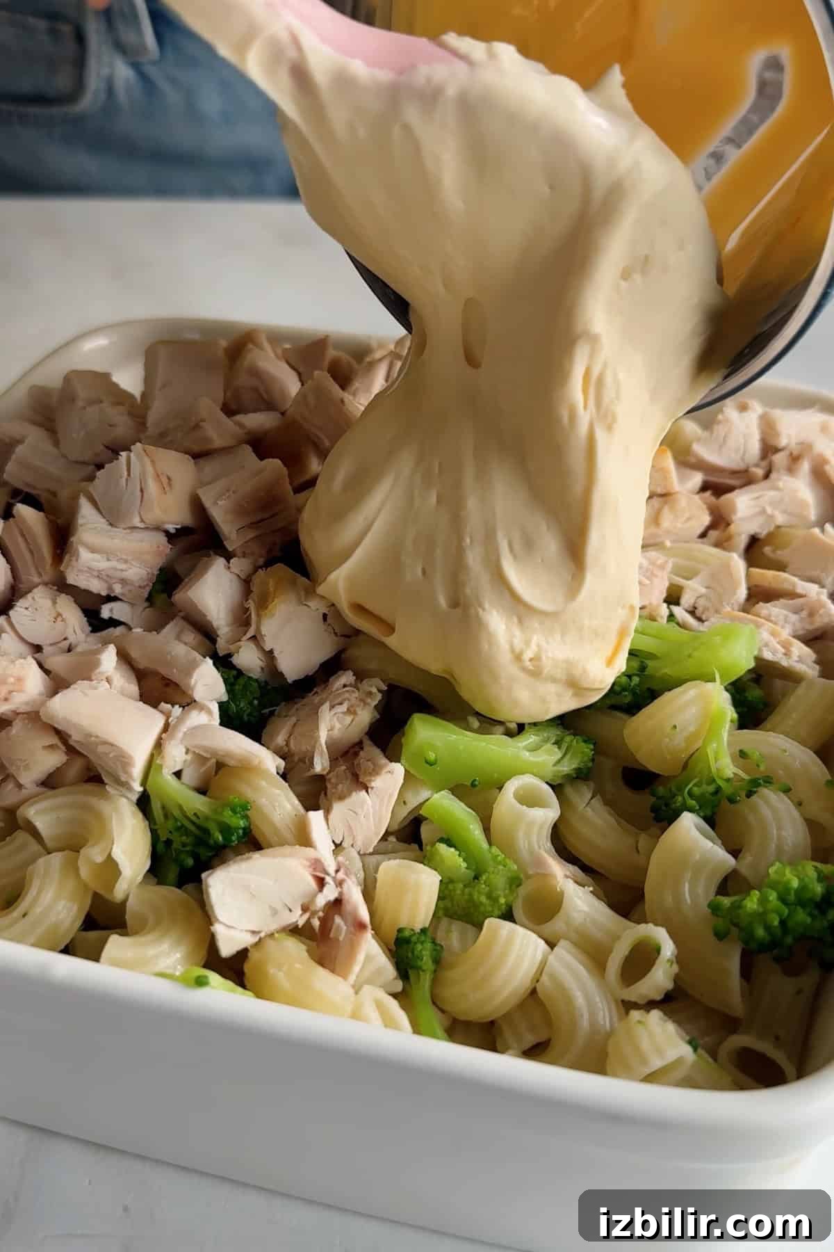 Creamy Chicken and Broccoli Mac with Cottage Cheese 4 Cheese sauce is being poured over chopped chicken, broccoli, and pasta in a white baking dish.