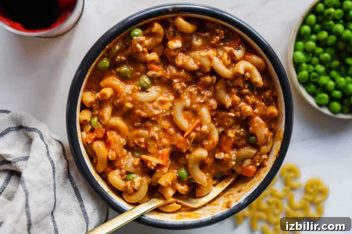 Creamy Cottage Cheese and Ground Beef Casserole 2 A bowl of macaroni in a thick tomato-based sauce with ground meat, peas, and carrots, with a spoon inside. A bowl of green peas and some dry macaroni are nearby.