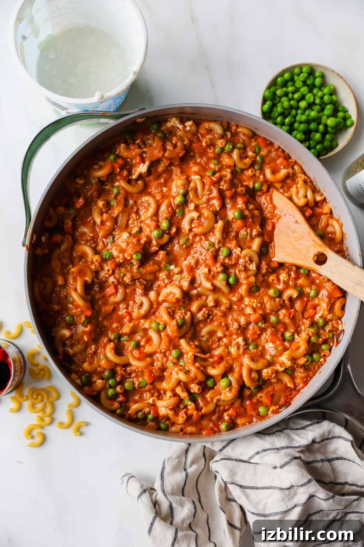 Creamy Cottage Cheese and Ground Beef Casserole 3 A pan of macaroni in tomato sauce with peas is being stirred with a wooden spoon. A bowl of peas, some dry macaroni, and a striped towel are nearby on a white surface.