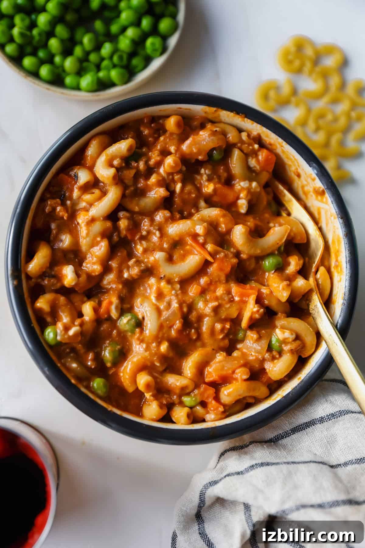 Creamy Cottage Cheese and Ground Beef Casserole 4 A bowl of macaroni in a thick tomato-based sauce with ground meat, peas, and carrots, with a spoon inside. A bowl of green peas and some dry macaroni are nearby.