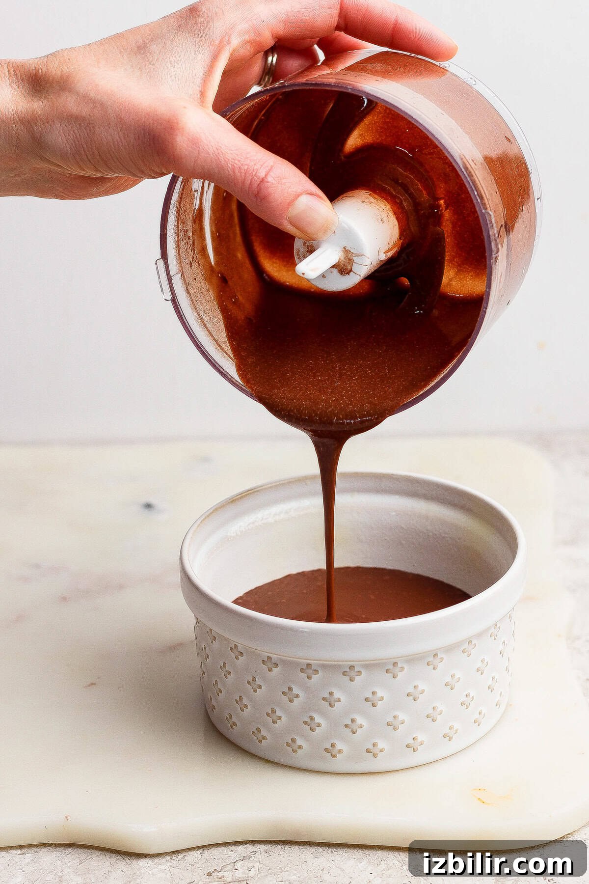 Single Serve Cottage Cheese Brownie Recipe for One 5 A hand pours thick chocolate sauce from a small food processor bowl into a white ceramic ramekin on a light surface.