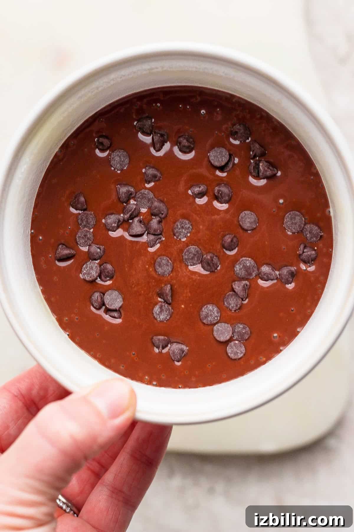 Single Serve Cottage Cheese Brownie Recipe for One 6 A hand holds a white bowl filled with chocolate batter topped with scattered chocolate chips.