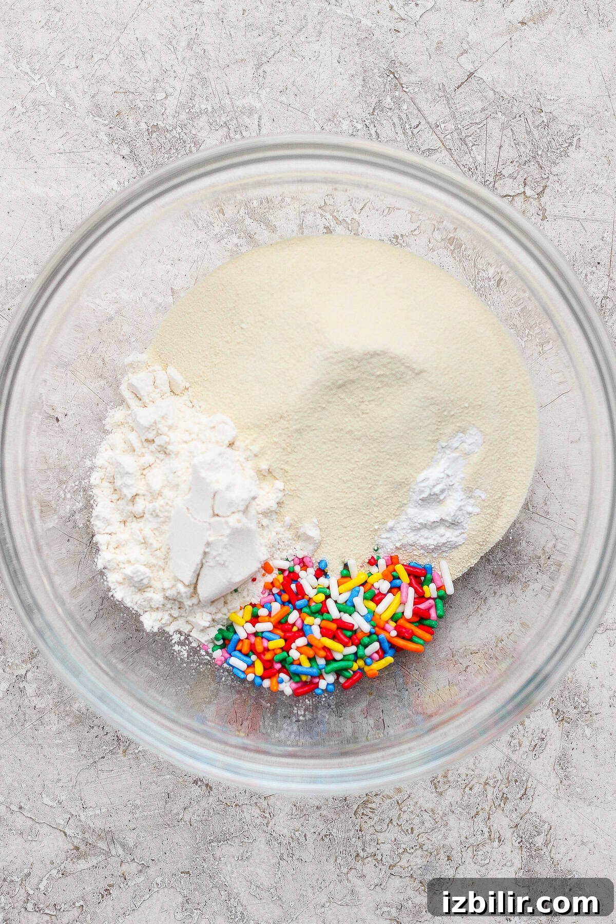 A glass bowl containing white flour, granulated sugar, baking powder, and colorful rainbow sprinkles on a light countertop.