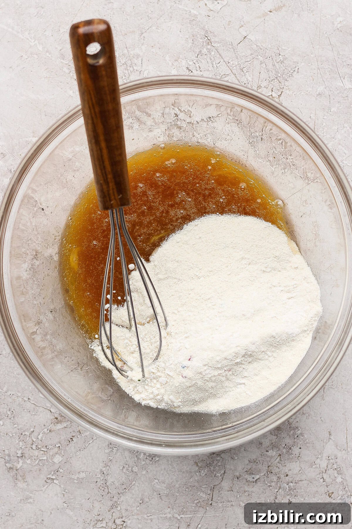 A glass bowl containing flour and a brown liquid being mixed with a metal whisk on a light textured surface.